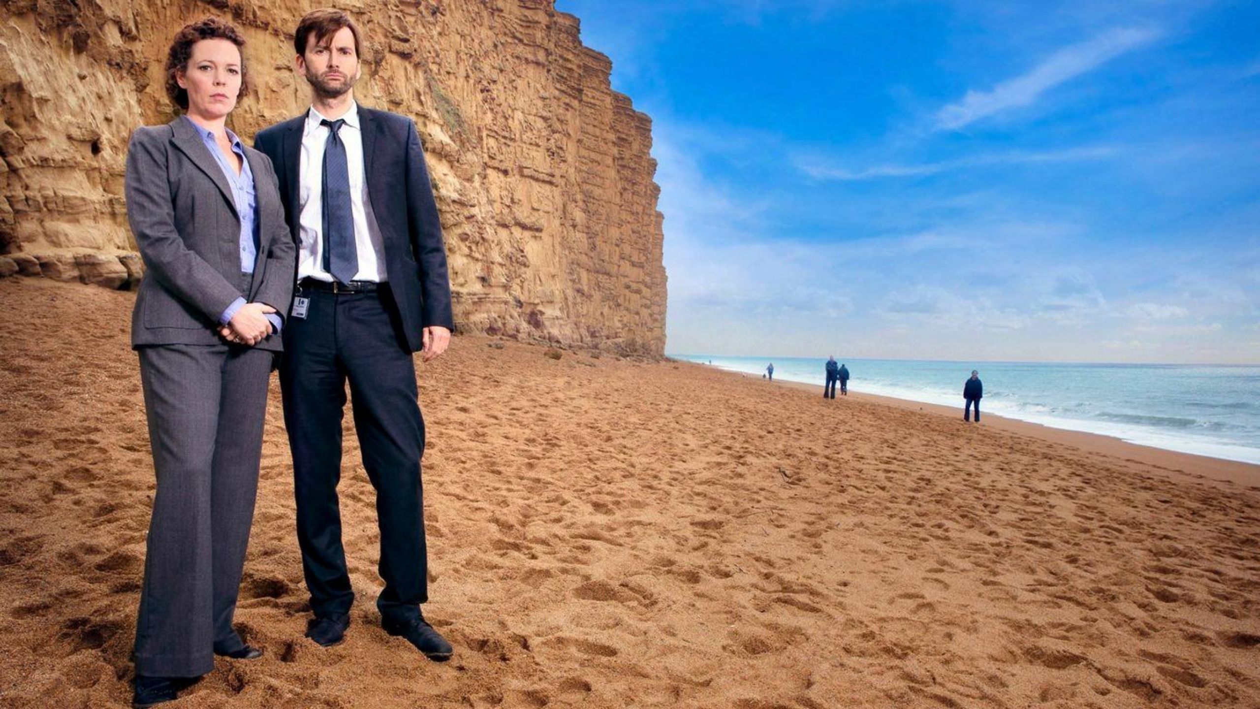 Olivia Colman and David Tenant stand in suits on a picturesque beach